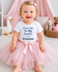 A smiling baby girl wearing a white t-shirt with text and a pink tutu sits on a white carpet, surrounded by stuffed animals and a pink canopy.