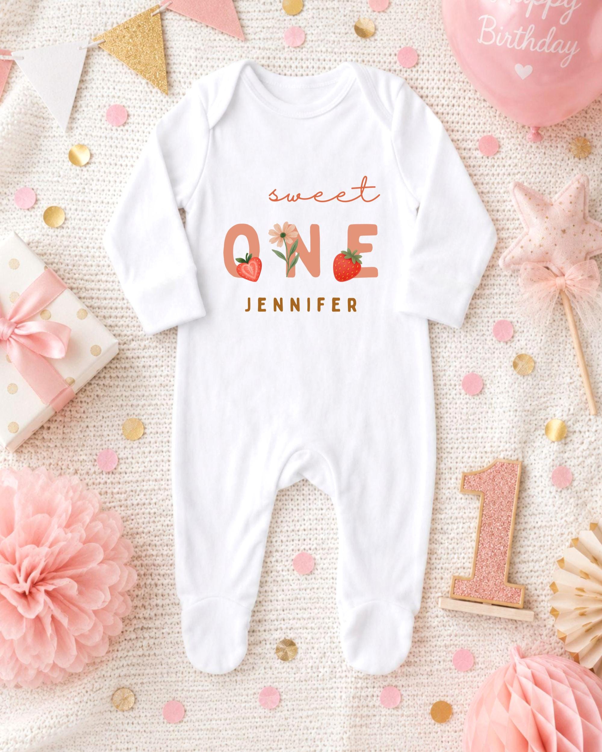 A white onesie with the text "Sweet One" and " Jennifer" printed on it, surrounded by pink and gold decorations, a pink star, and a pink balloon.