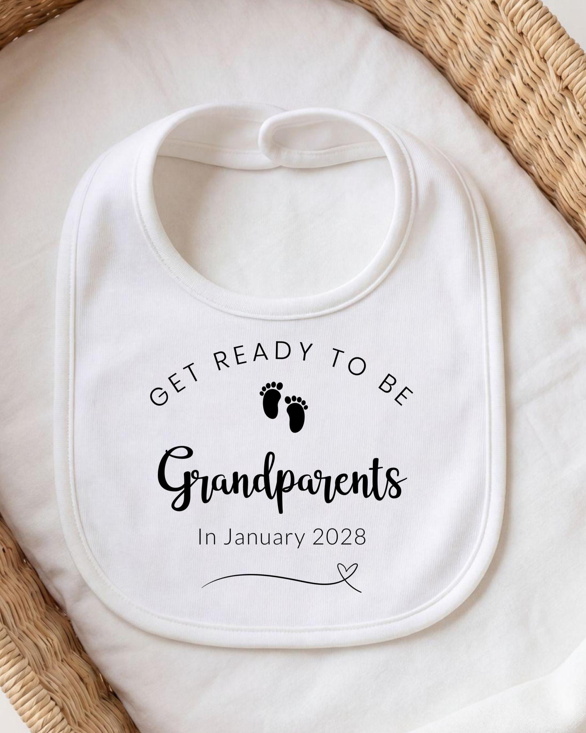 A white bib with the text "GET READY TO BE Grandparents In January 2028" printed on it, along with a small graphic of baby feet.