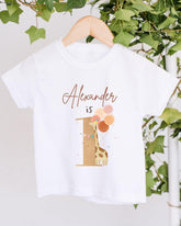 A white t-shirt with a giraffe and balloons printed on it, hanging on a wooden hanger.