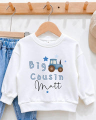 A white sweatshirt with a tractor design and the text "Big Cousin Matt" printed on it, hanging on a wooden hanger.