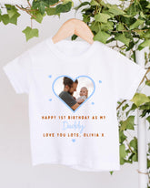A white t-shirt with a photo of a man holding a baby inside a heart shape, along with the text "Happy 1st Birthday as my Daddy Love you lots, Olivia X".
