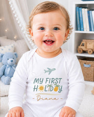 A smiling baby wearing a white onesie with the text "My first holiday - Diana" printed on it, sitting in a room with a teddy bear and bookshelf in the background.