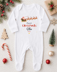 A white onesie with a Christmas-themed design, including a Santa Claus sleigh and reindeer, is displayed on a white background alongside various Christmas decorations and ornaments.