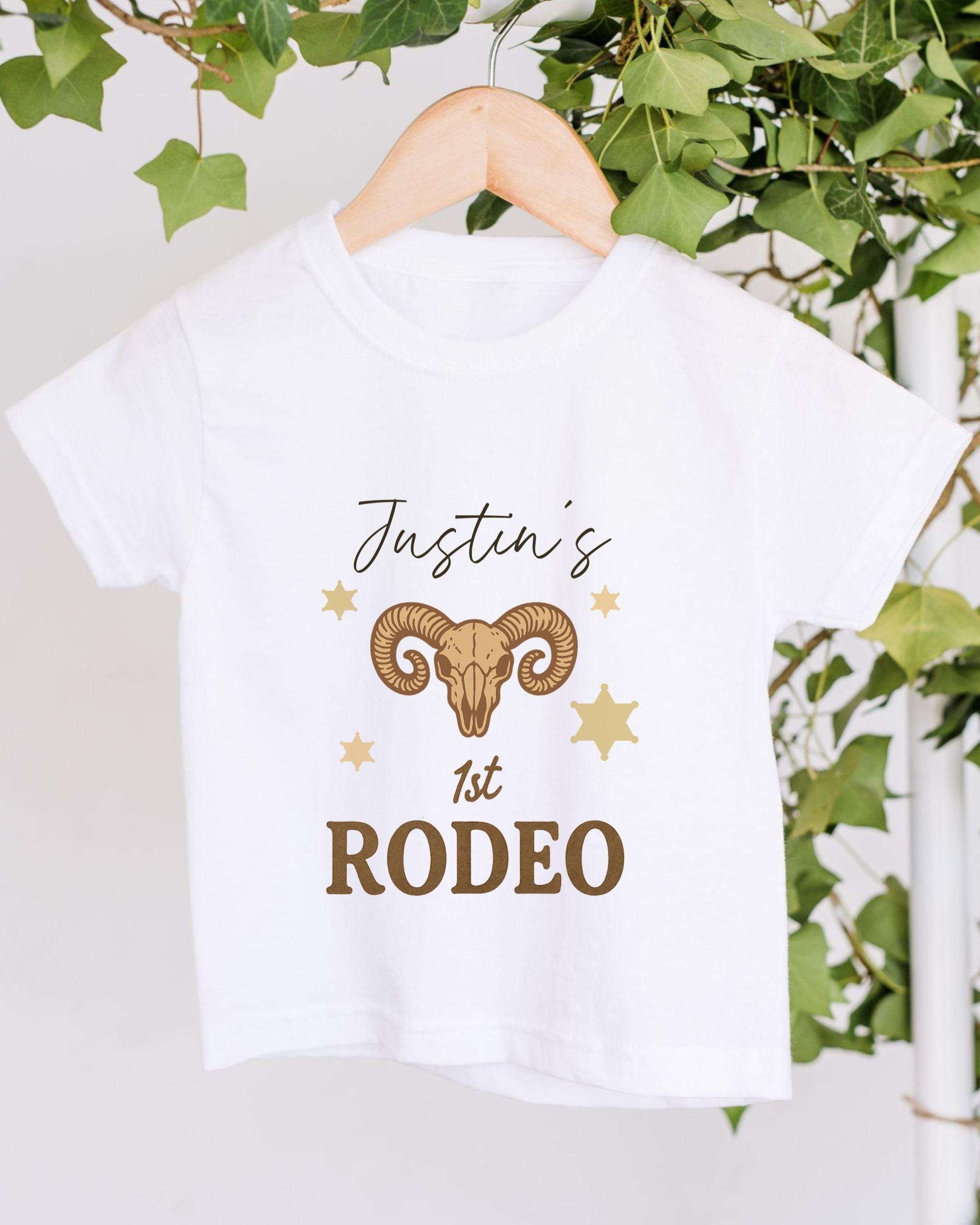 A white t-shirt with a graphic design featuring a ram's head and stars, along with the text "Justin's 1st Rodeo".