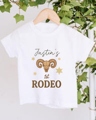 A white t-shirt with a graphic design featuring a ram's head and stars, along with the text "Justin's 1st Rodeo".