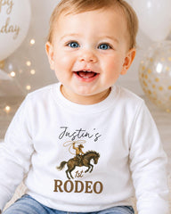 A smiling baby boy wearing a white shirt with a cowboy graphic and the text " Justin's 1st Rodeo" printed on it.