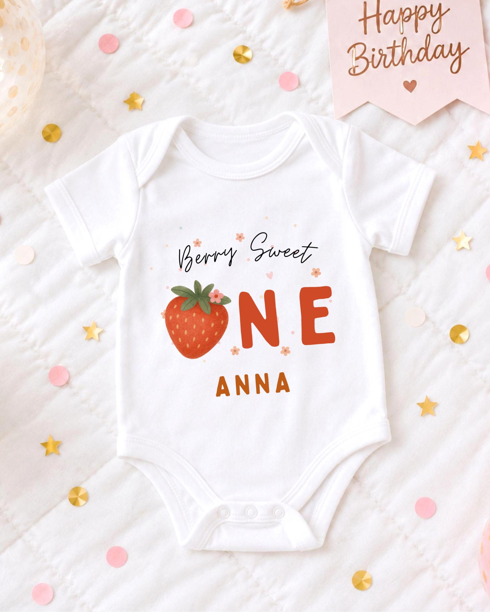 A white onesie with a strawberry design and the text "Berry Sweet One Anna" printed on it, placed on a bed with confetti and a birthday card.