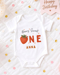 A white onesie with a strawberry design and the text "Berry Sweet One Anna" printed on it, placed on a bed with confetti and a birthday card.