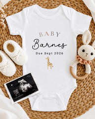 A white baby onesie with the text "BABY Barnes Due Sept 2026" printed on it, along with a stuffed animal and a pair of crocheted baby booties. There is also an Ultrasound image of a baby's belly.