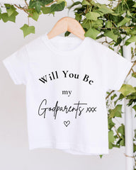 A white t-shirt with the text "Will You Be my Godparents xxx" printed on it, hanging on a wooden hanger.