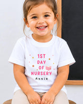 A young girl wearing a white t-shirt with the text "1st DAY OF NURSERY" and "Annie" printed on it, smiling at the camera.