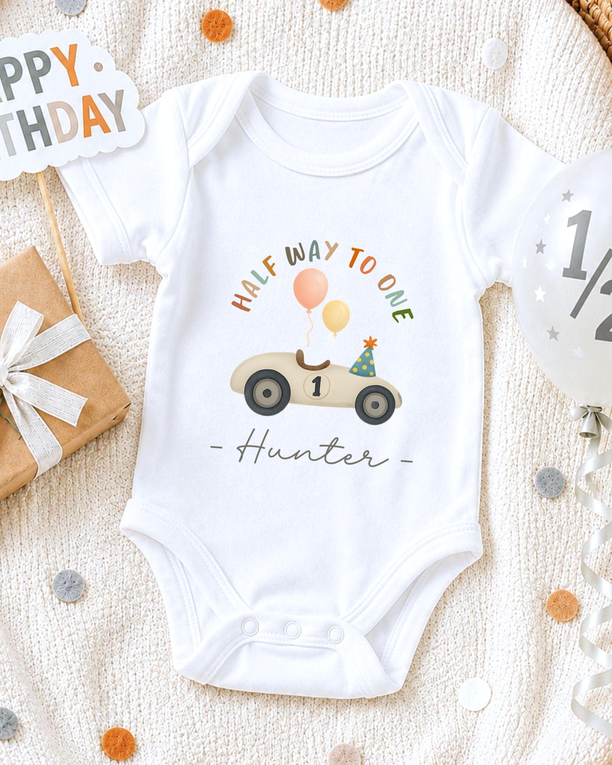 A white onesie with a cartoon car and balloons, along with a "Happy Birthday Hunter" message, is displayed on a bed with confetti.