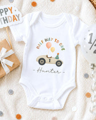 A white onesie with a cartoon car and balloons, along with a "Happy Birthday Hunter" message, is displayed on a bed with confetti.