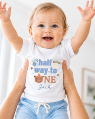 A smiling baby wearing a white onesie with a birthday message and a cartoon character.