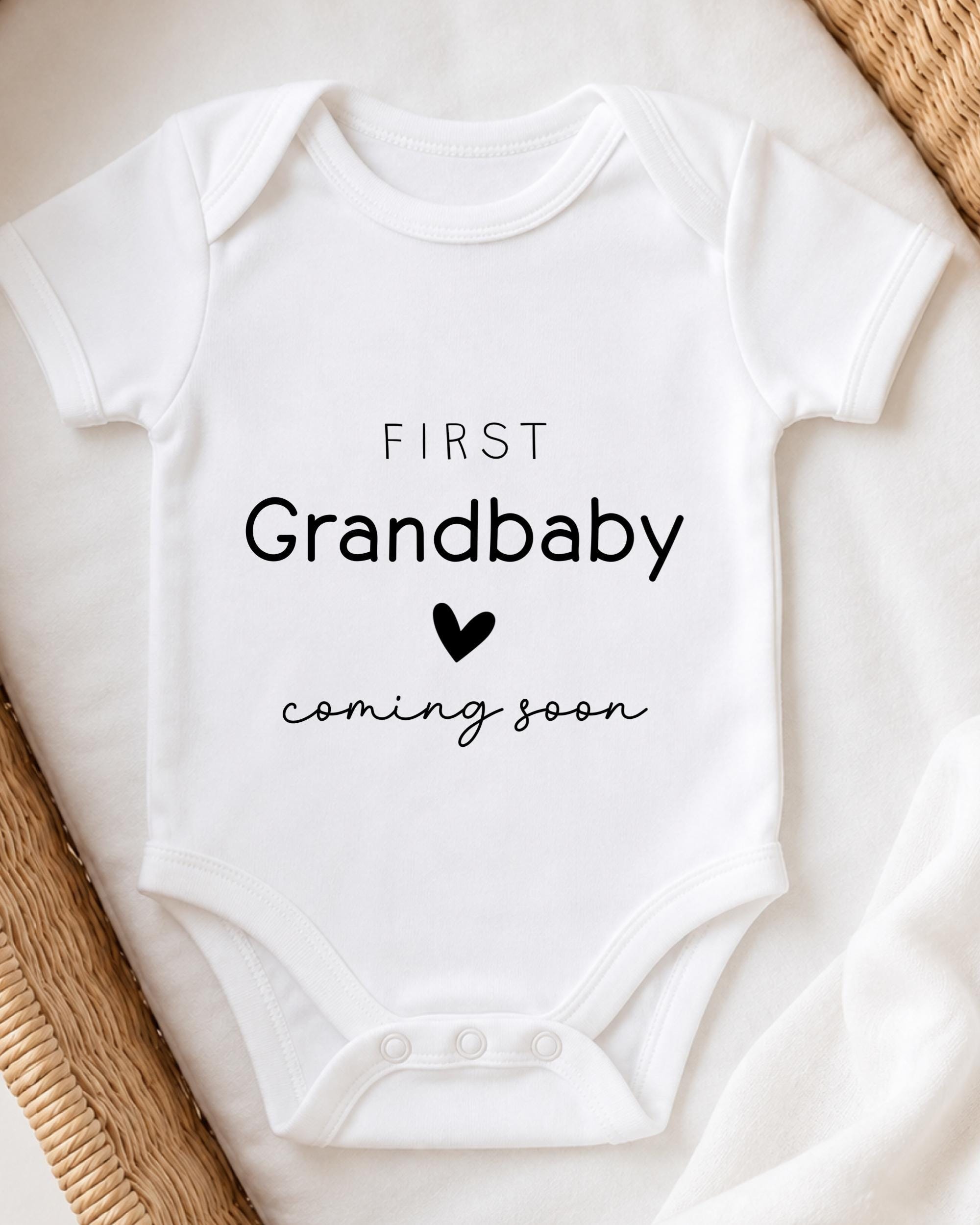 A white onesie with the text "FIRST Grandbaby coming soon" printed on it.