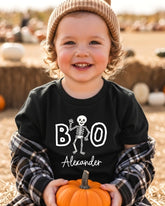 A young child wearing a black t-shirt with a skeleton design and the name "Alexander" printed on it, holding an orange pumpkin.