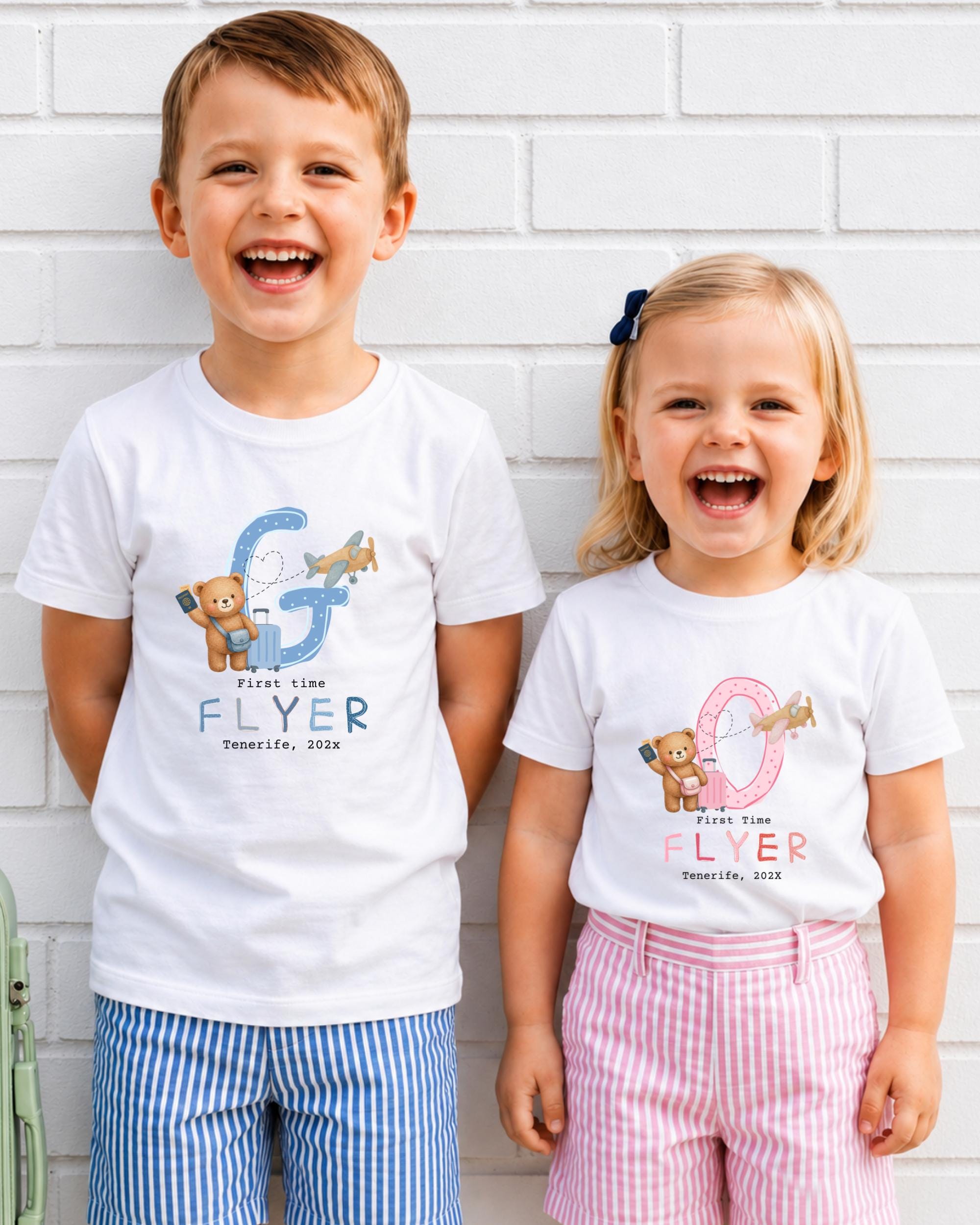 Two children, a boy and a girl, are standing next to each other and smiling. They are both wearing white t-shirts with a graphic design featuring a letter 'G' and a teddy bear.