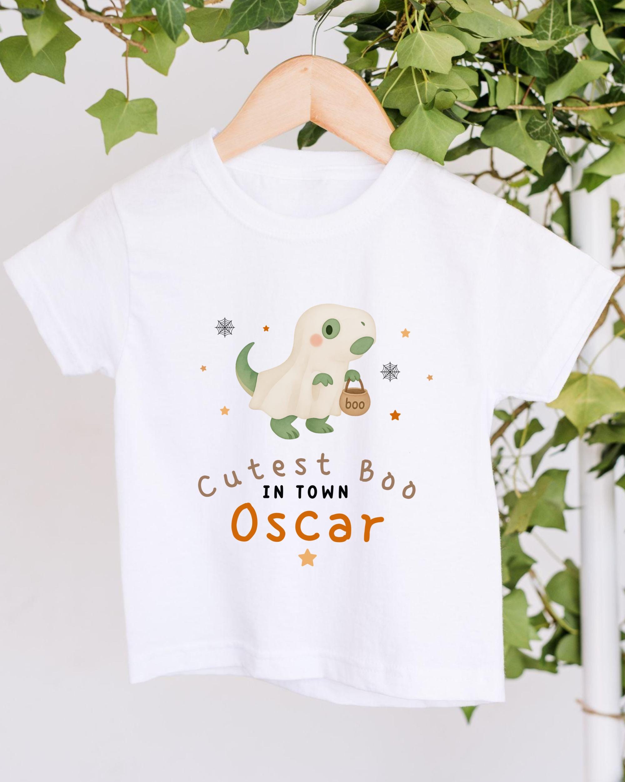 A white t-shirt with a cartoon dinosaur and the text "Cute in town Oscar" printed on it, hanging on a wooden hanger.