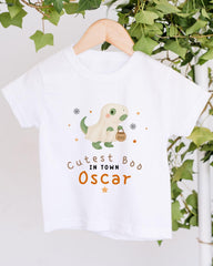 A white t-shirt with a cartoon dinosaur and the text "Cute in town Oscar" printed on it, hanging on a wooden hanger.