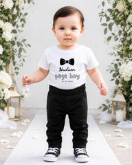 A young child wearing a white t-shirt with a bow tie and black pants stands on a white carpet in front of a floral archway.
