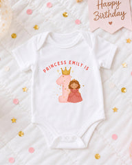 A white onesie with a princess and a crown, along with the text "PRINCESS EMILY IS" and a number 1, is displayed on a bed with confetti and a birthday card.