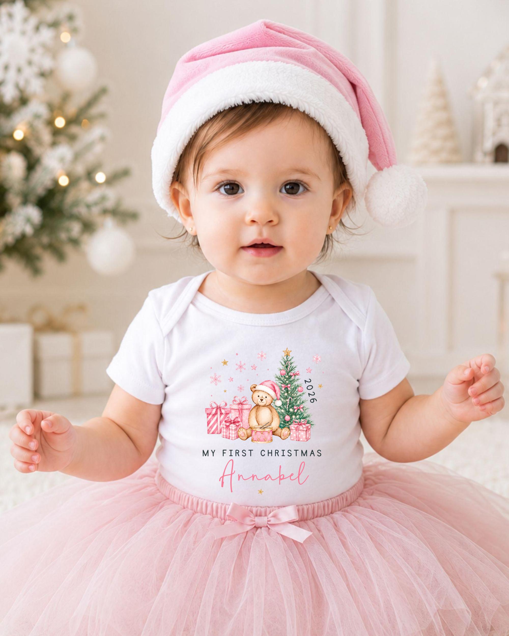 A young girl wearing a pink Santa hat and a white t-shirt with a Christmas-themed design, sitting on a white carpet in front of a Christmas tree.