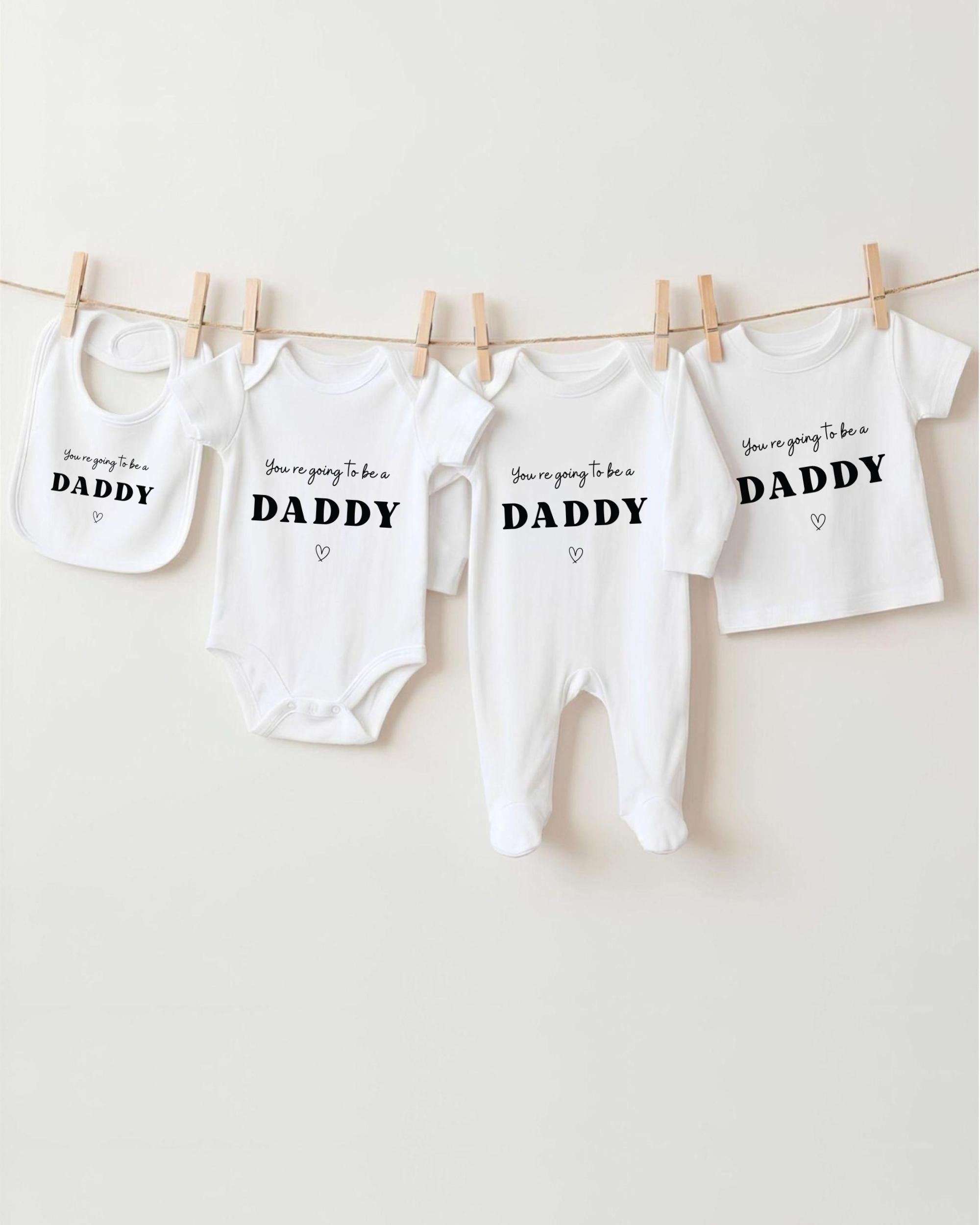 A row of four white baby clothes with the text "You're going to be a DADDY" printed on them, hanging on a clothesline.