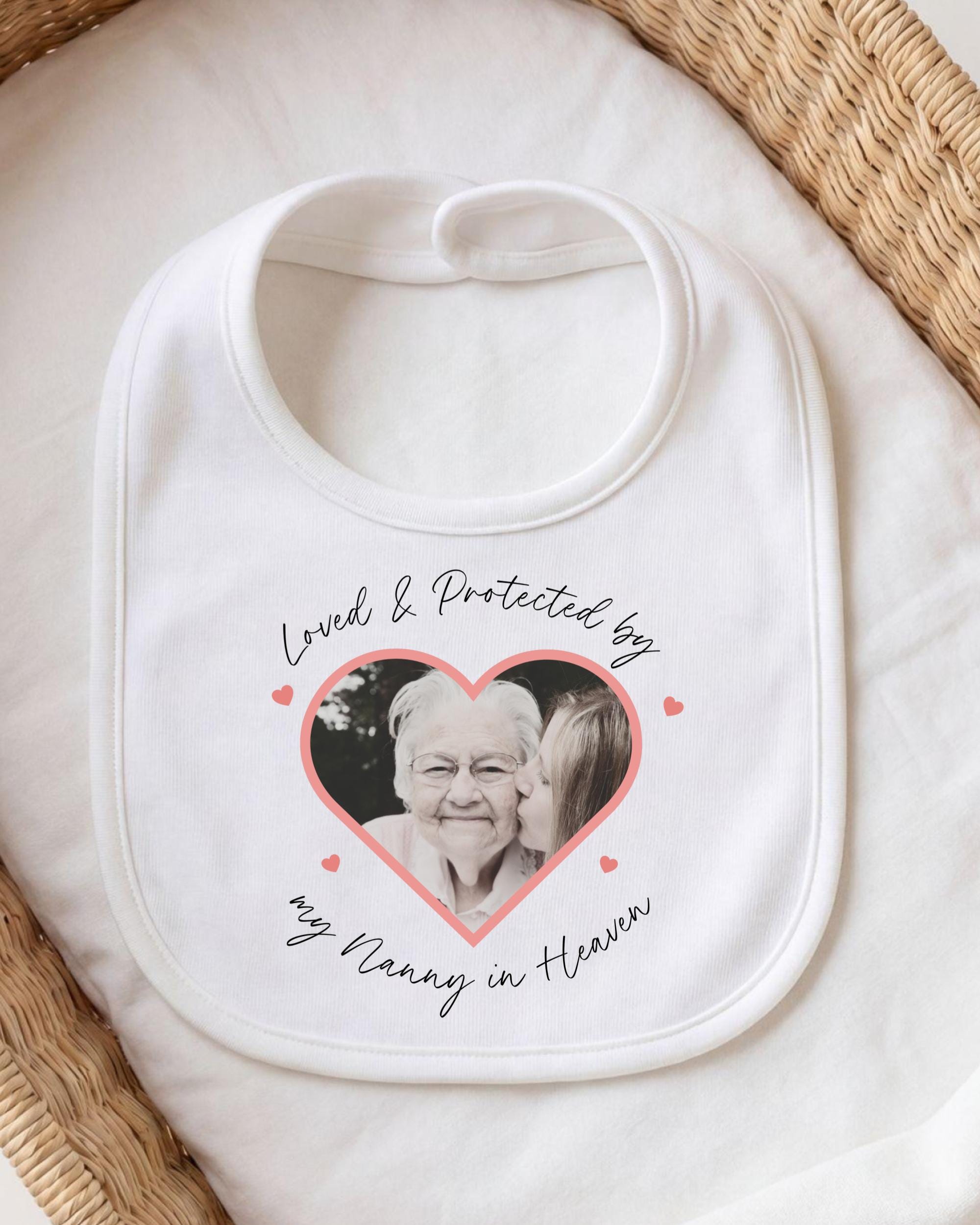 A white bib with a heart-shaped photo of an elderly woman and the text "Loved & Protected by my Nanny in Heaven" printed on it.