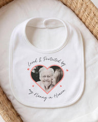A white bib with a heart-shaped photo of an elderly woman and the text "Loved & Protected by my Nanny in Heaven" printed on it.