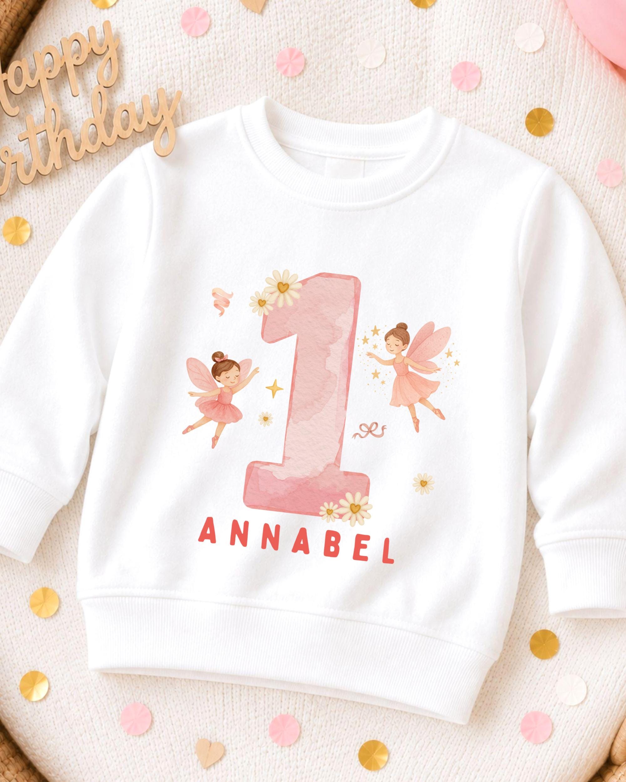A white sweatshirt with a pink number one and two-tailed fairies printed on it, along with the name 'Annabel' written below.
