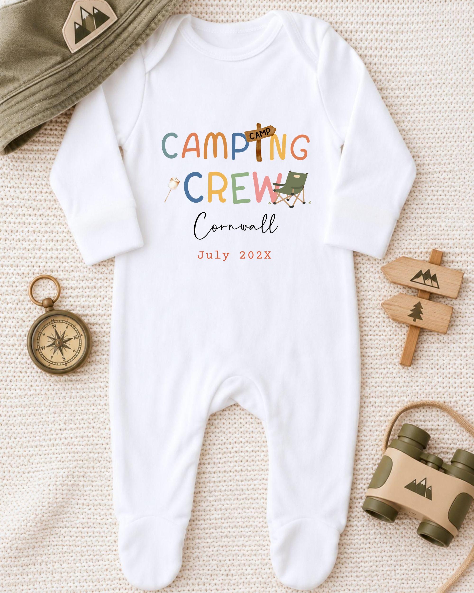 A baby onesie with the text "Camping Crew Cornwall July 2023" printed on it, along with camping-related items such as a hat, a compass, and a pair of binoculars.