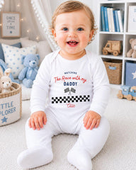 A smiling baby boy wearing a white onesie with a racing theme, sitting on the floor in a nursery setting with various toys and books around him.