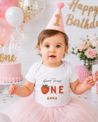 A young girl wearing a white t-shirt with a strawberry design and a pink party hat sits in front of a birthday cake and balloons.