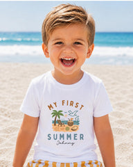 A young boy with a big smile on his face, wearing a white t-shirt with a graphic design of a beach scene and the text "My first summer" printed on it.