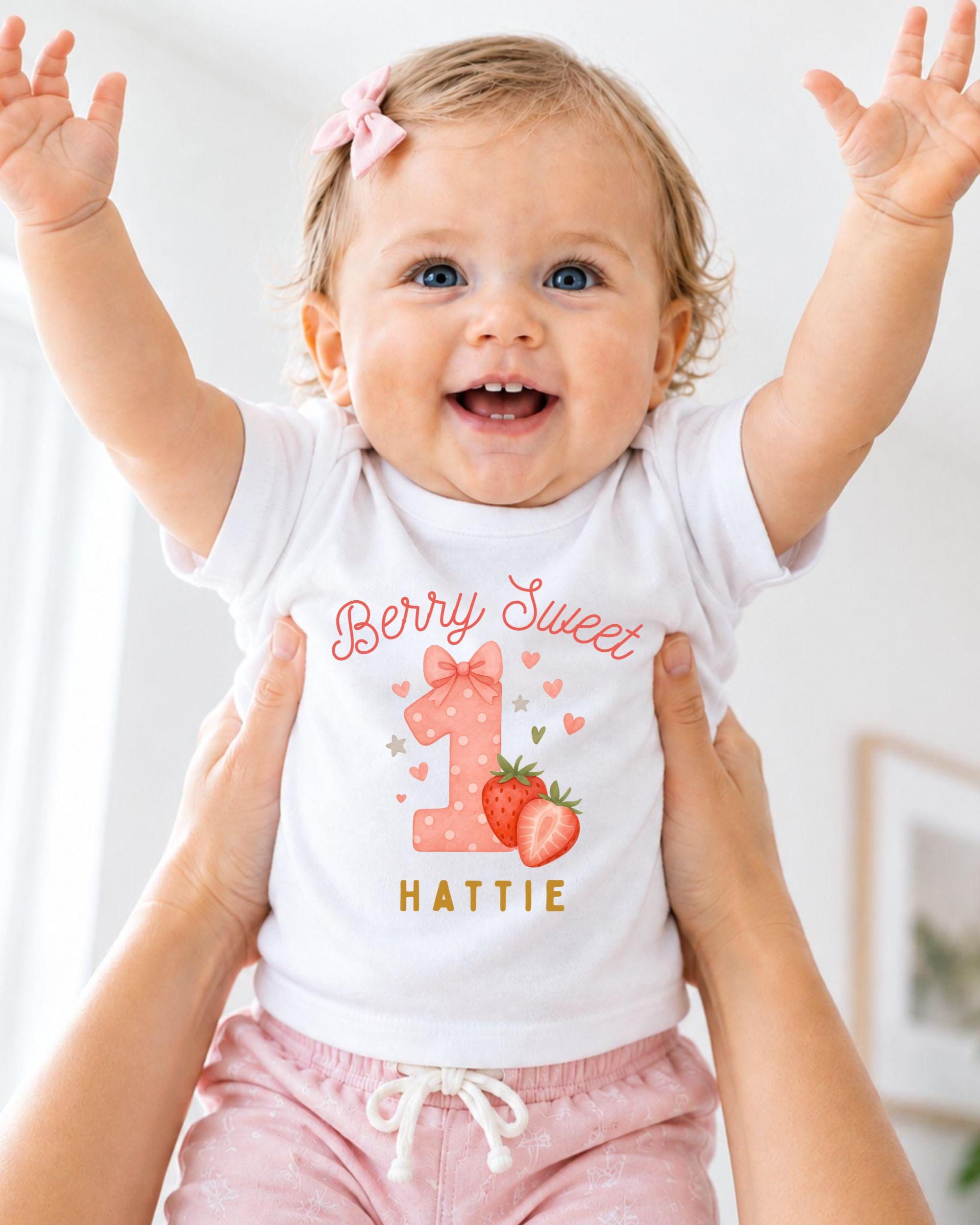 A smiling baby girl wearing a white t-shirt with a personalized design and the text "Berry Sweet Hattie" is being held up by someone's arms.