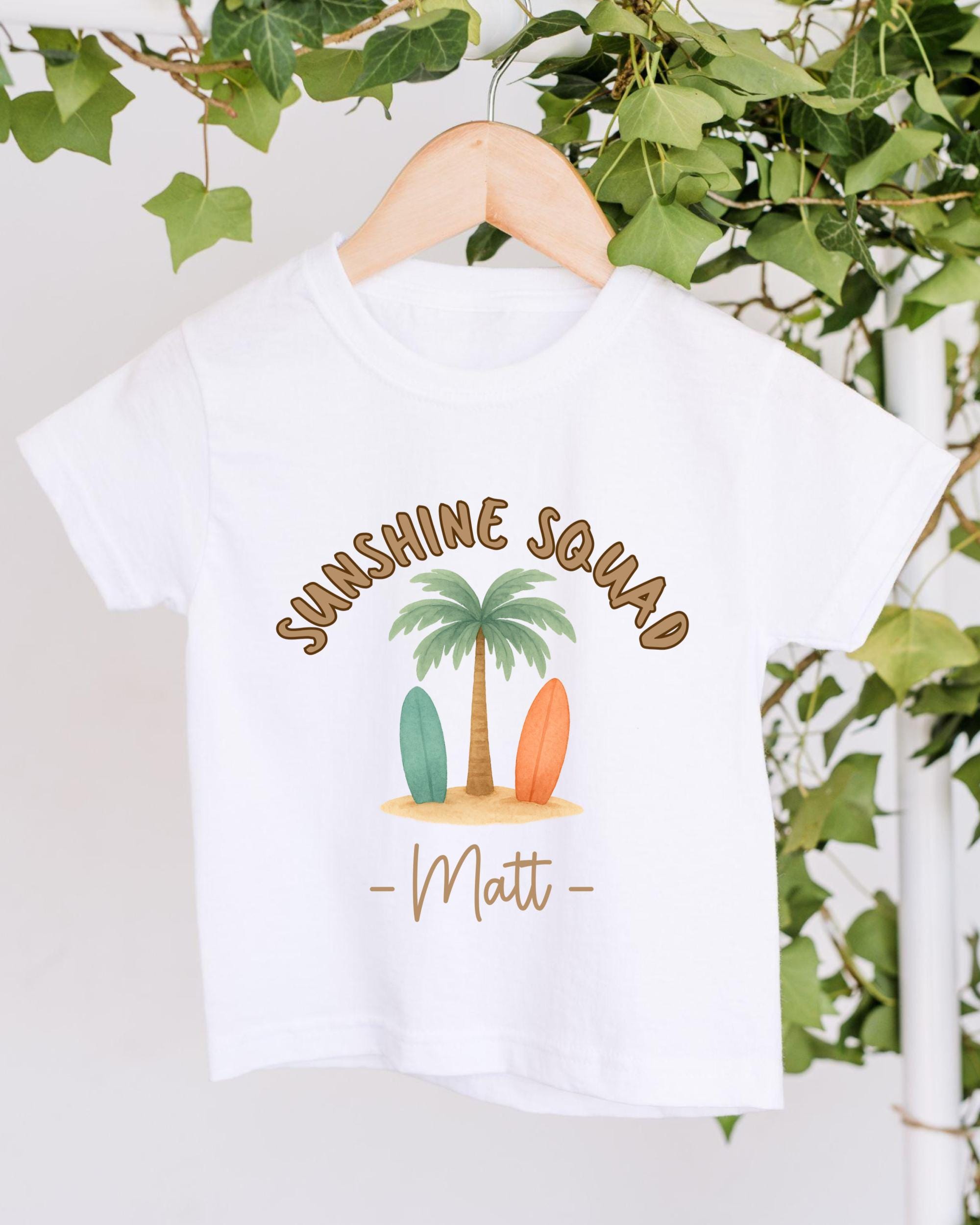 A white t-shirt with a graphic design featuring a palm tree, two surfboards, and the text "SUNSHINE SQUAD Matt" printed on it.