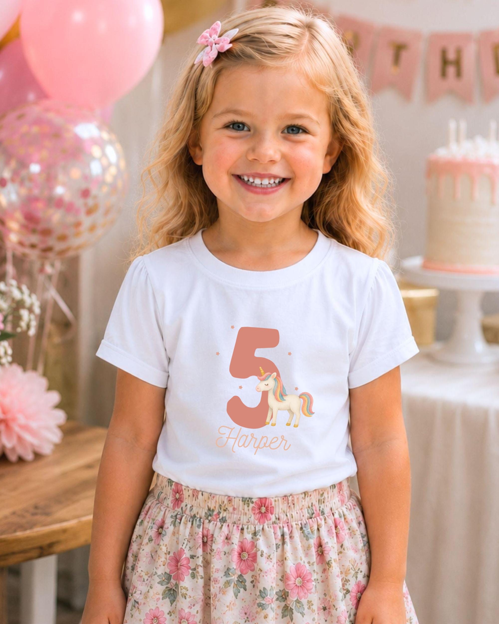 A young girl wearing a white t-shirt with a unicorn graphic and the number 5, smiling at the camera.