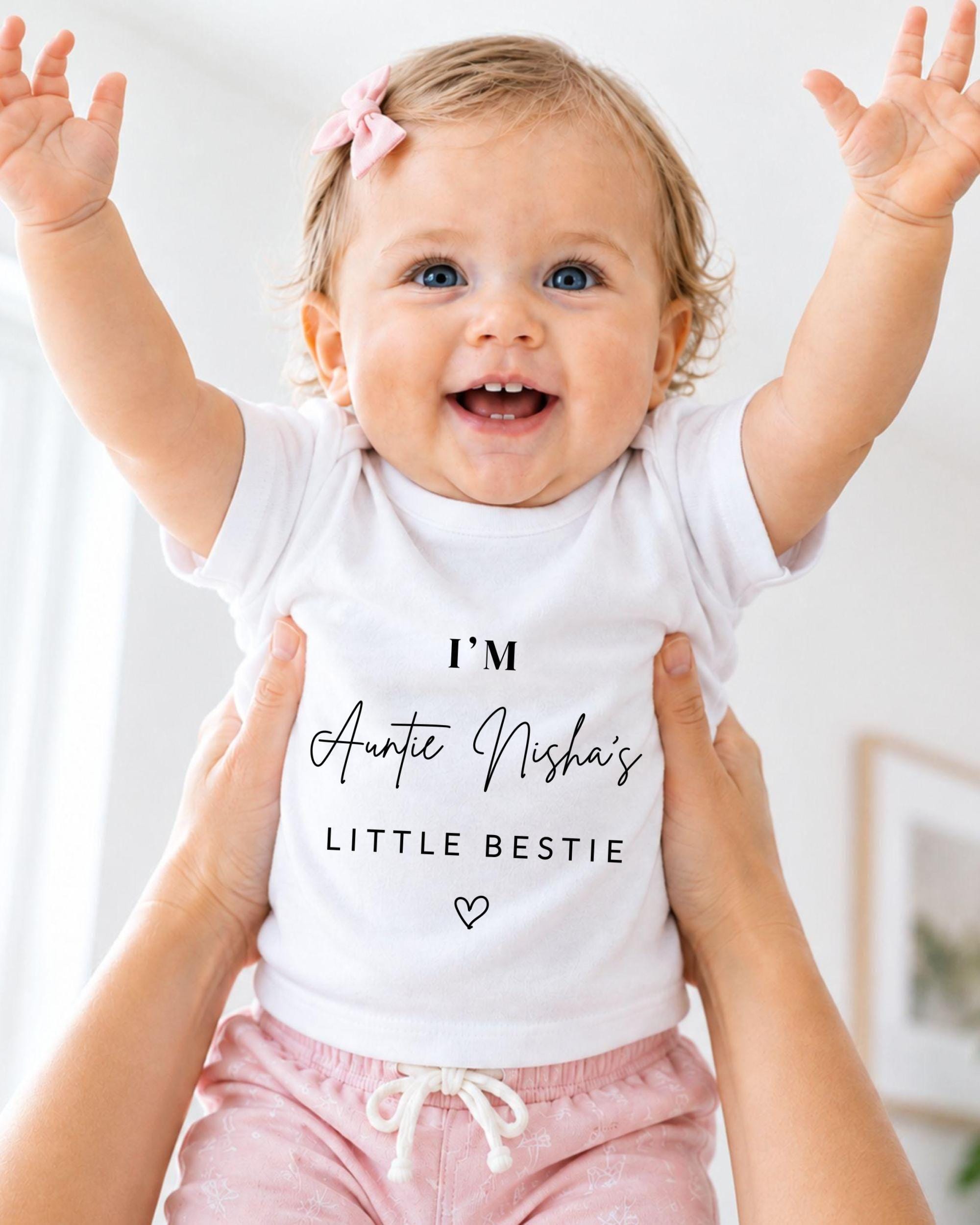 A smiling baby girl wearing a white t-shirt with text and a heart symbol, being held up by someone's arms.