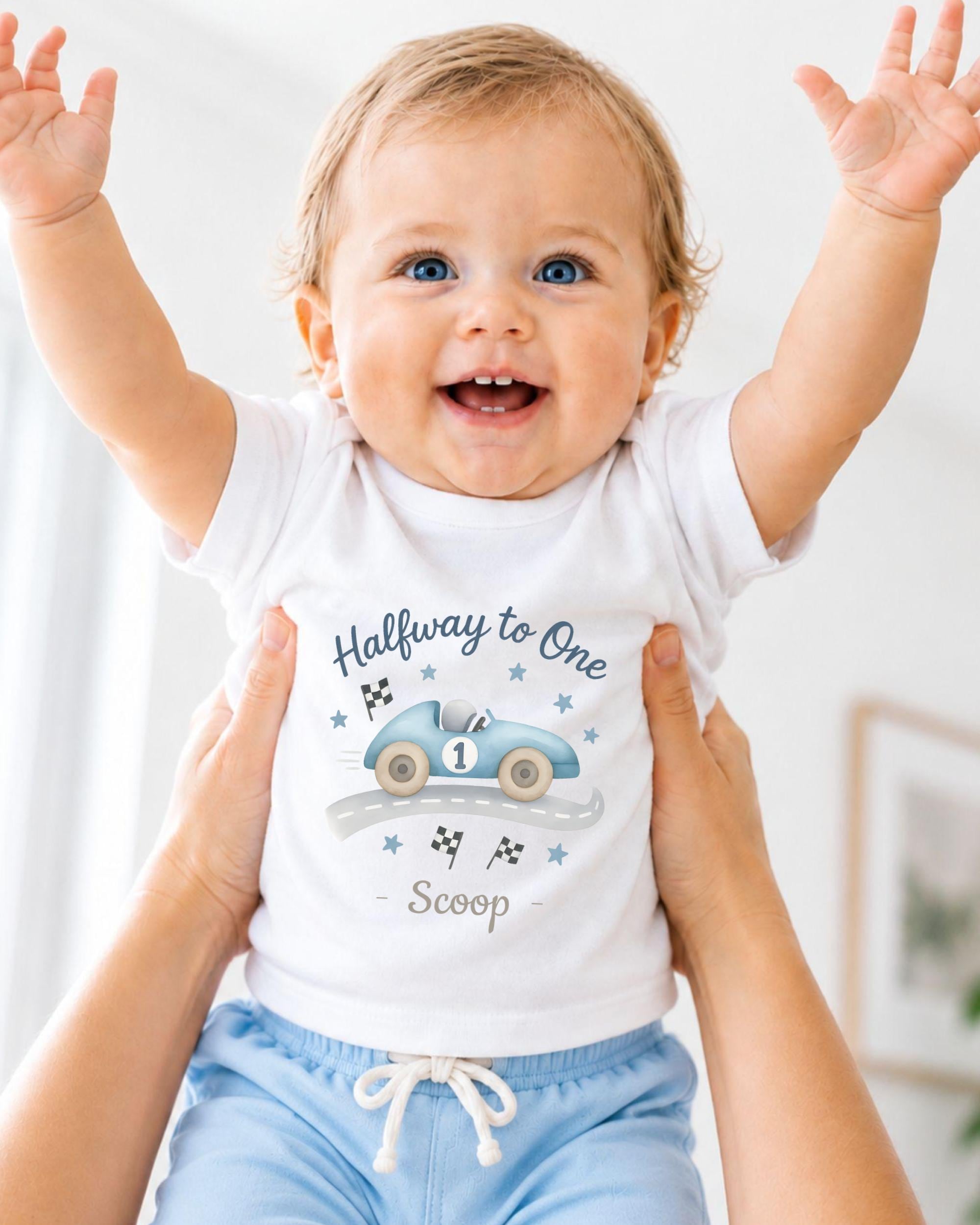 A smiling baby wearing a white t-shirt with a race car graphic and the text "Halfway to One Scoop" is being held up by someone's arms.