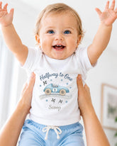 A smiling baby wearing a white t-shirt with a race car graphic and the text "Halfway to One Scoop" is being held up by someone's arms.