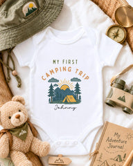a baby onesie with a camping trip design, surrounded by camping-related items such as a hat, compass, binoculars, and a teddy bear.
