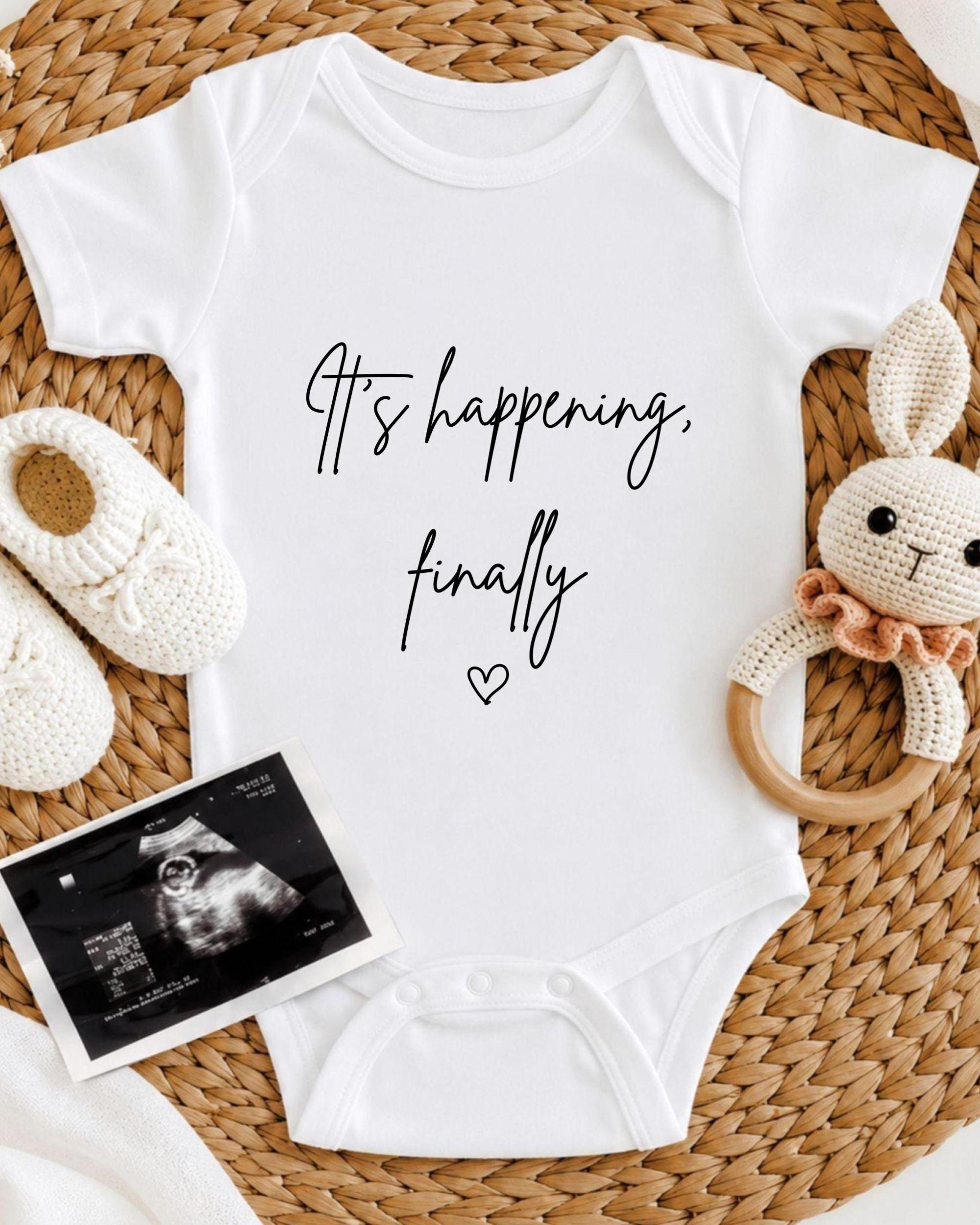 A white baby onesie with the text "It's happening, finally" printed on it, along with a crocheted bunny toy and a black and white photograph of an Ultrasound image.