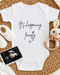 A white baby onesie with the text "It's happening, finally" printed on it, along with a crocheted bunny toy and a black and white photograph of an Ultrasound image.