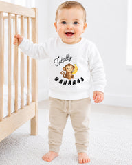 A smiling baby boy wearing a white sweatshirt with a monkey and the text "Totally Bananas" is standing next to a wooden crib.