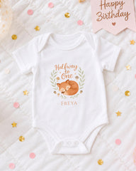A white onesie with a cartoon fox design and the text "Halfway to One" printed on it, placed on a bed with confetti and a "Happy Birthday" sign.