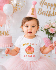 A young girl wearing a birthday hat and a white t-shirt with a strawberry donut graphic is posing in front of a birthday cake and balloons.