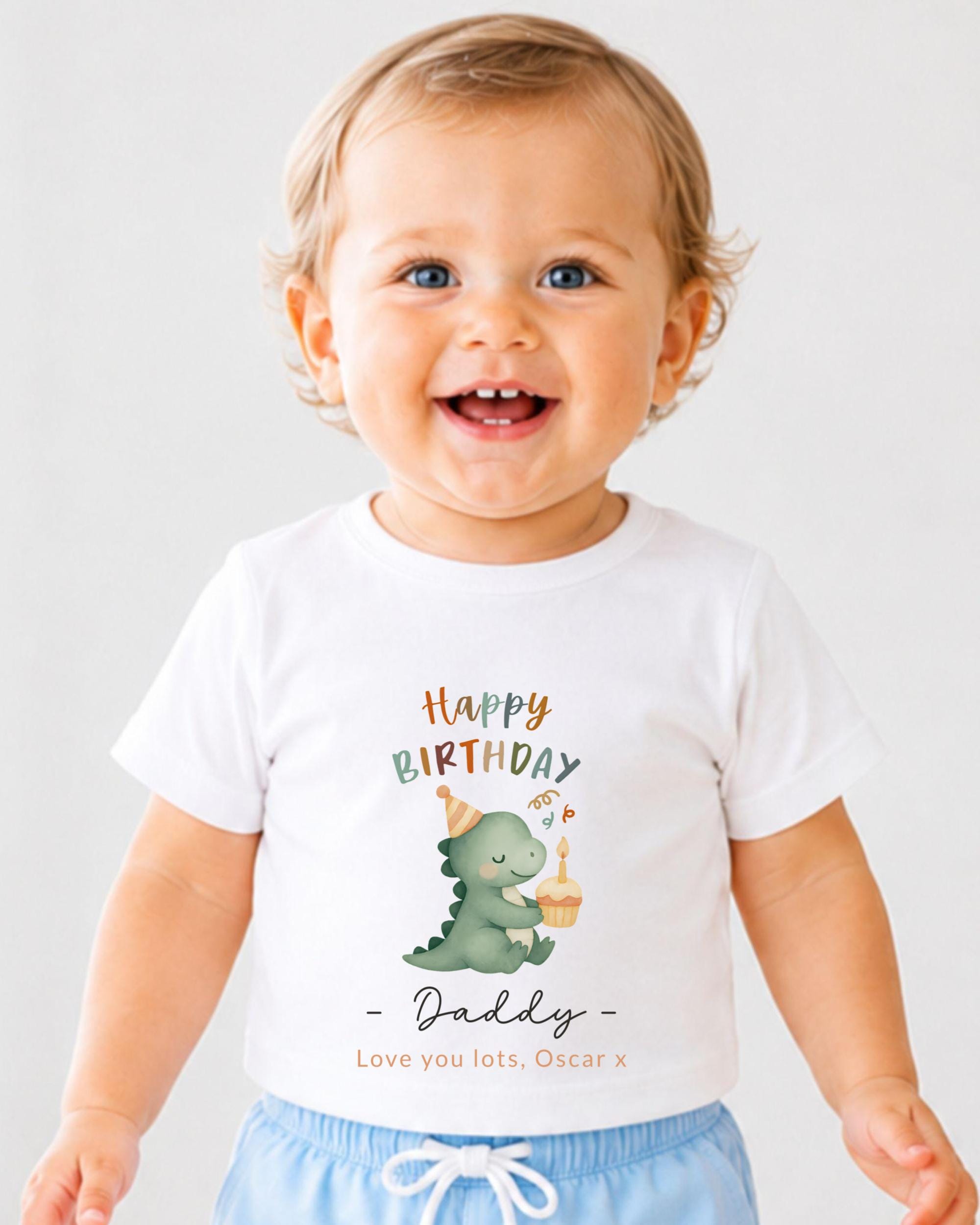 A smiling baby boy wearing a white t-shirt with a dinosaur graphic and text that says "Happy Birthday Daddy - Love you lots, Oscar X".