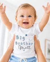 A smiling baby wearing a white shirt with the text "Big Brother Matt" and blue shorts, being held up by someone's arms.
