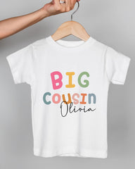 A person's hand is holding a white t-shirt with the text "BIG COUSIN Olivia" printed on it.